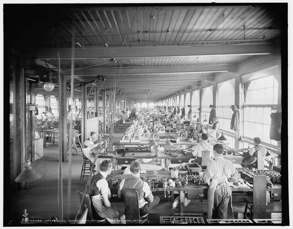 Black and white photograph of the assembling department at the National Cash Register factory in Dayton, Ohio, around 1902, with rows of workers at benches.